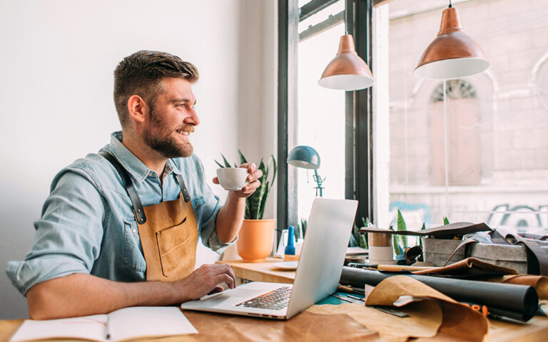 Profile view of a handsome man sitting in his workshop, using his laptop, drinking coffee, smiling